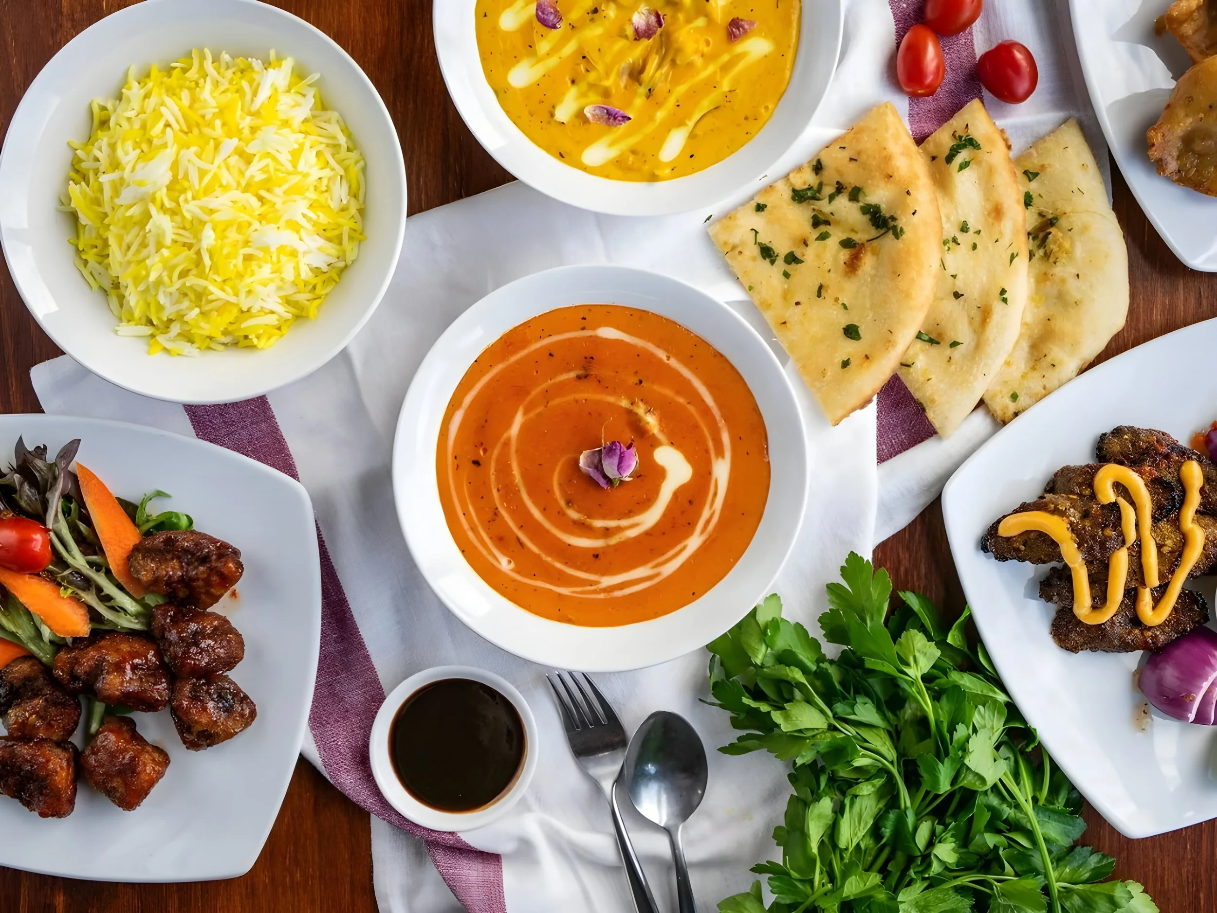 Assorted dishes of Indian cuisine on a wooden table, including rice, curry, naan bread, salad, and various sauces, garnished with herbs and vegetables.