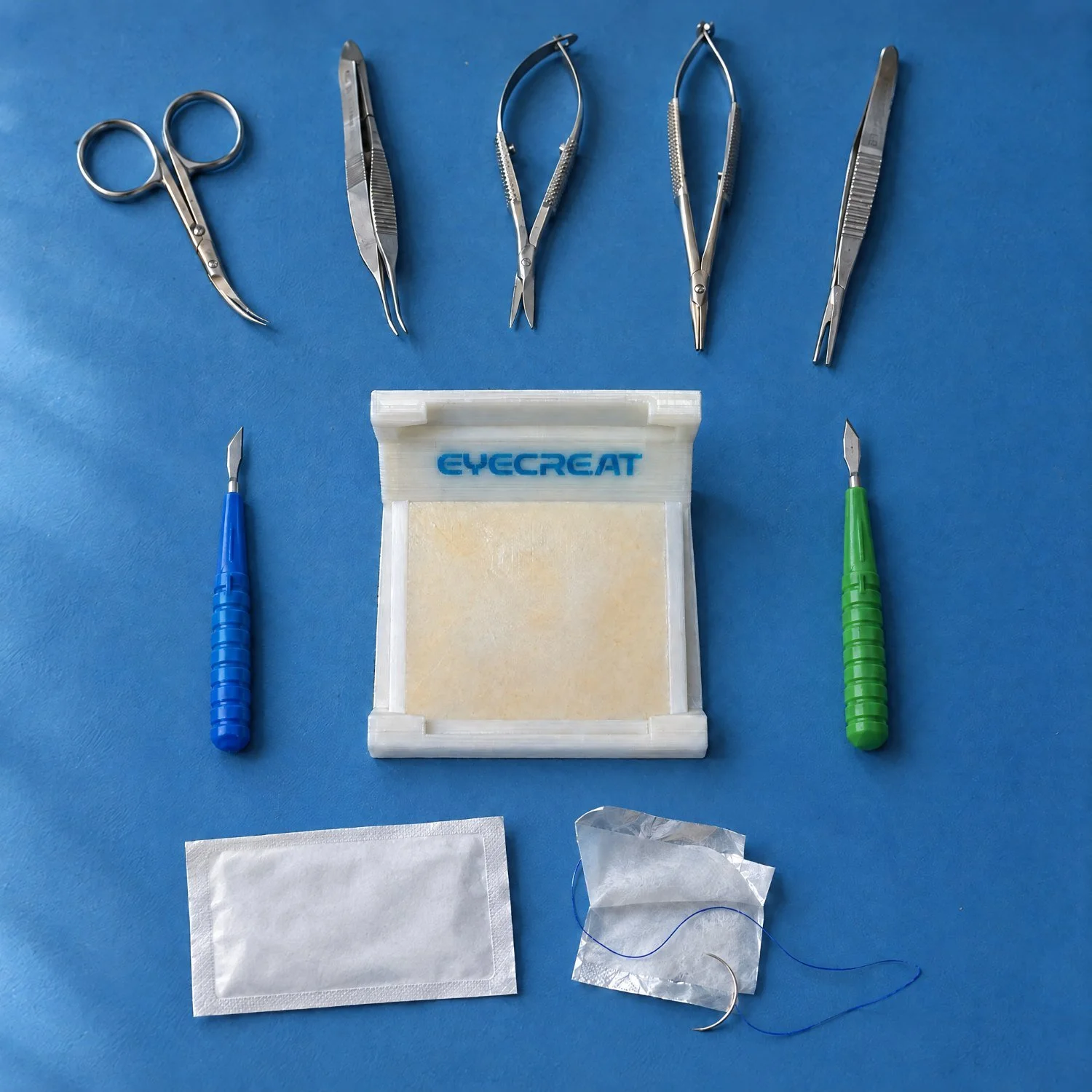 Kit 4 Conjunctival Suturing laid out on a blue cloth: Holder 4 ConjSuturing with pad, conjunctival scissors, forceps, Westcott scissors, needle holder, ligature forceps, crescent and 15 degree knife, and two absorbable sutures. Training products by eyecre.at.