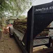 A trailer filled with logs parked outdoors, with trees and a house nearby.