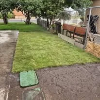 A backyard with a small grassy area, a concrete path, a wooden bench, and a fenced yard with trees.