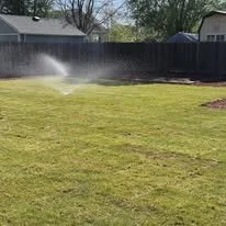 A backyard with a well-maintained grassy lawn and a sprinkler watering the grass, with a fence and some trees in the background.