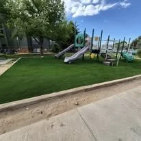 Children's playground with slide and climbing equipment on artificial grass near a sidewalk and trees.