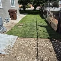 A backyard with new sod being installed, showing a section of freshly laid grass and another section of bare soil and gravel.