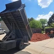 A dump truck unloading red soil onto a sidewalk in a residential area with trees and houses in the background.