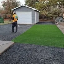 A person standing near a newly installed section of green artificial turf in a backyard with a garage and trees in the background.