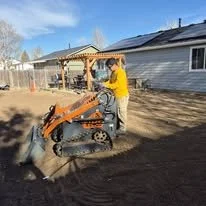 A person operating a small orange mini excavator in a residential backyard during daytime.