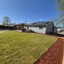 A house with a large backyard, green grass, and a dirt border, with trees and a clear blue sky in the background.