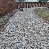 A gravel pathway bordered by bricks, with grass on one side and a wooden fence in the background.