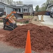 A pile of red mulch in a residential driveway with a small skid-steer loader nearby, and an orange traffic cone in the foreground.