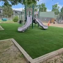 A playground with two gray slides, surrounded by green grass and trees.