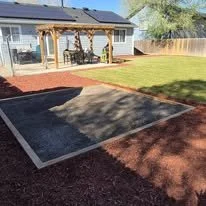 Backyard with a partially shaded trampoline area, surrounded by red brick and garden grass, with a wooden patio and house in the background.