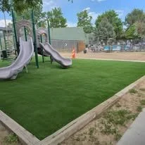 Playground with two slides and artificial grass area, surrounded by a wooden border, in a park.