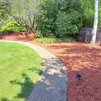 A curved garden pathway with grass on one side and mulched garden beds on the other, surrounded by trees and bushes.