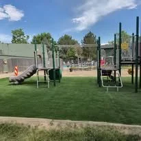 Playground with slides and climbing equipment on a grassy field, with trees and a fence in the background under a blue sky.