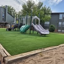 Playground with slides and climbing equipment on a grassy area, with a building in the background.