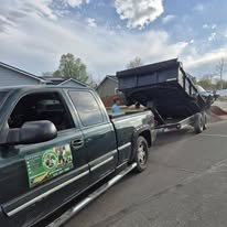 Green pickup truck with a logo on the door, parked beside a black dump trailer that is tipped forward, spilling debris onto the ground.