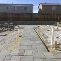 A freshly paved stone walkway leading towards a fenced backyard with houses in the background.