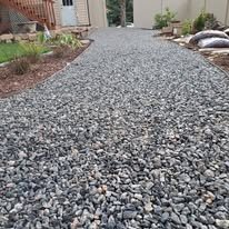 Gravel driveway with plants and rocks on the side, leading to a house in the background.