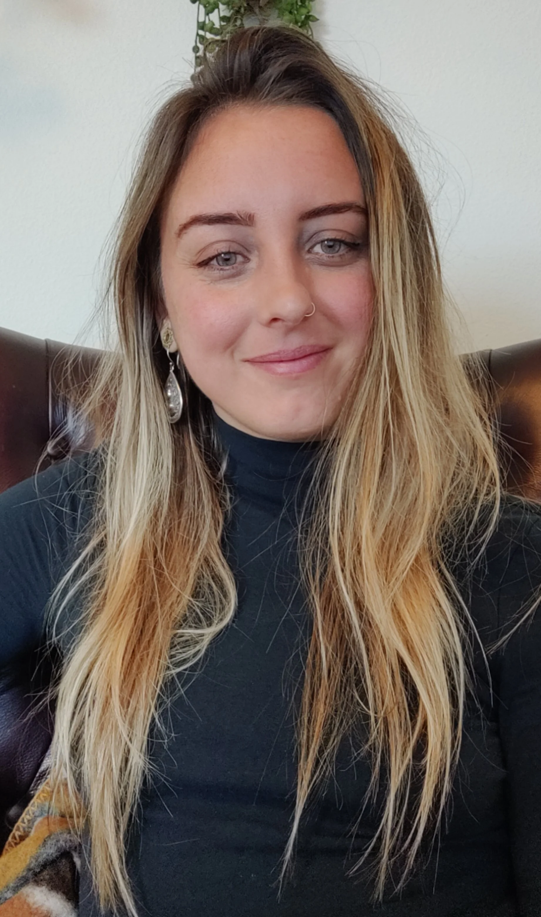A woman with long wavy hair and light-colored eyes, wearing earrings and a dark top, smiling while sitting on a brown leather couch with a real or faux plant in the background.