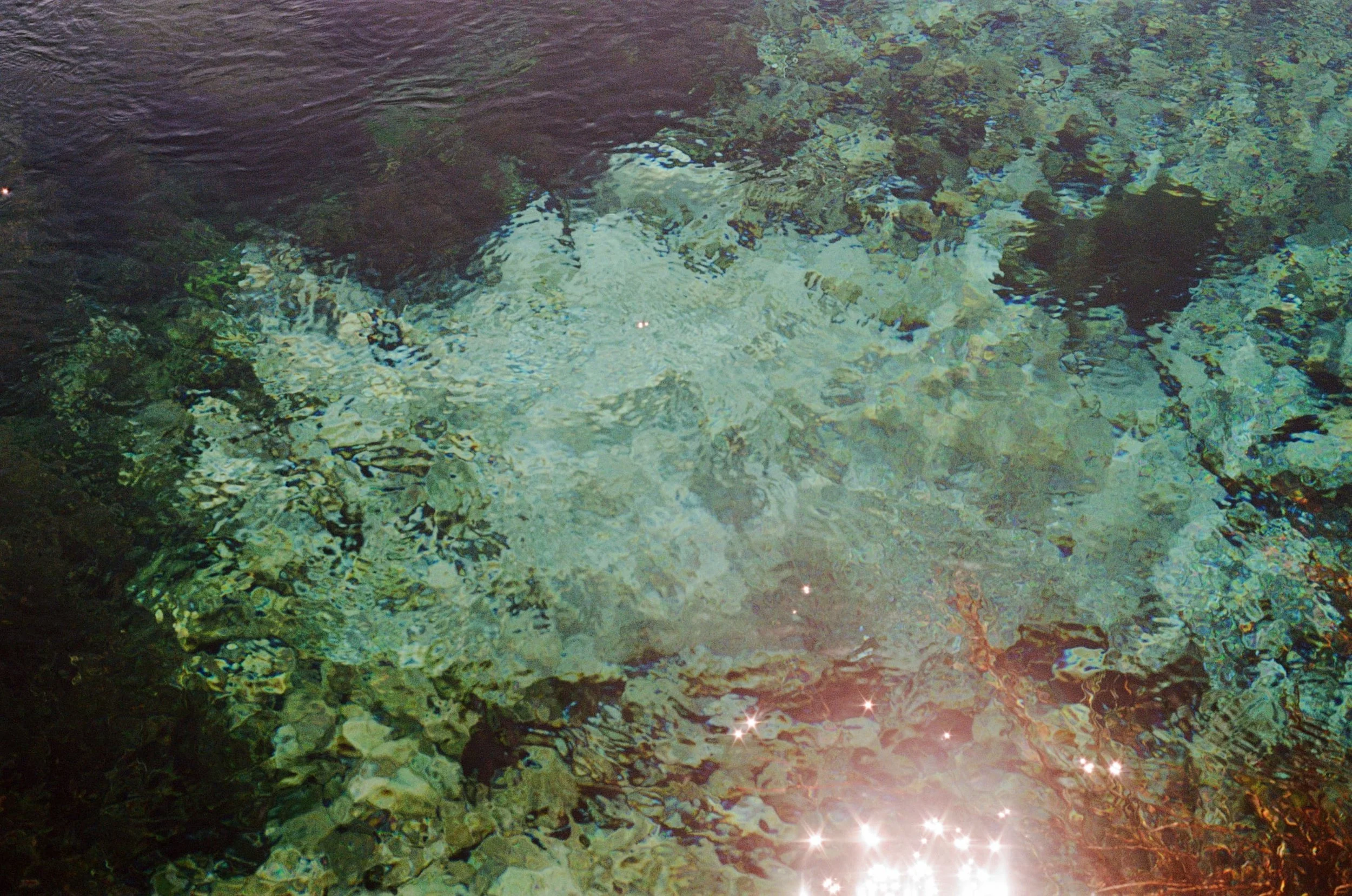 Clear water with rocks and pebbles visible at the bottom, sunlight reflecting on the surface, creating sparkle effects.
