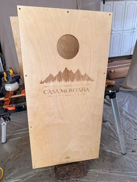 Finished cornhole board standing in the Lito WoodWorks garage workshop, showing the completed Casa Montaña laser engraving with mountain silhouette and decorative typography.