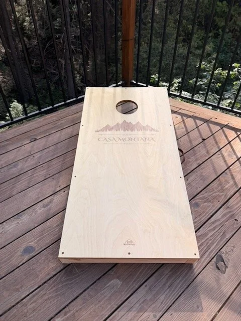 Regulation-size cornhole board built from 3/4" Baltic birch plywood, laser-engraved with a custom mountain silhouette design. Finished with oil-based Varathane and photographed at the Casa Montaña property in Placerville, California.