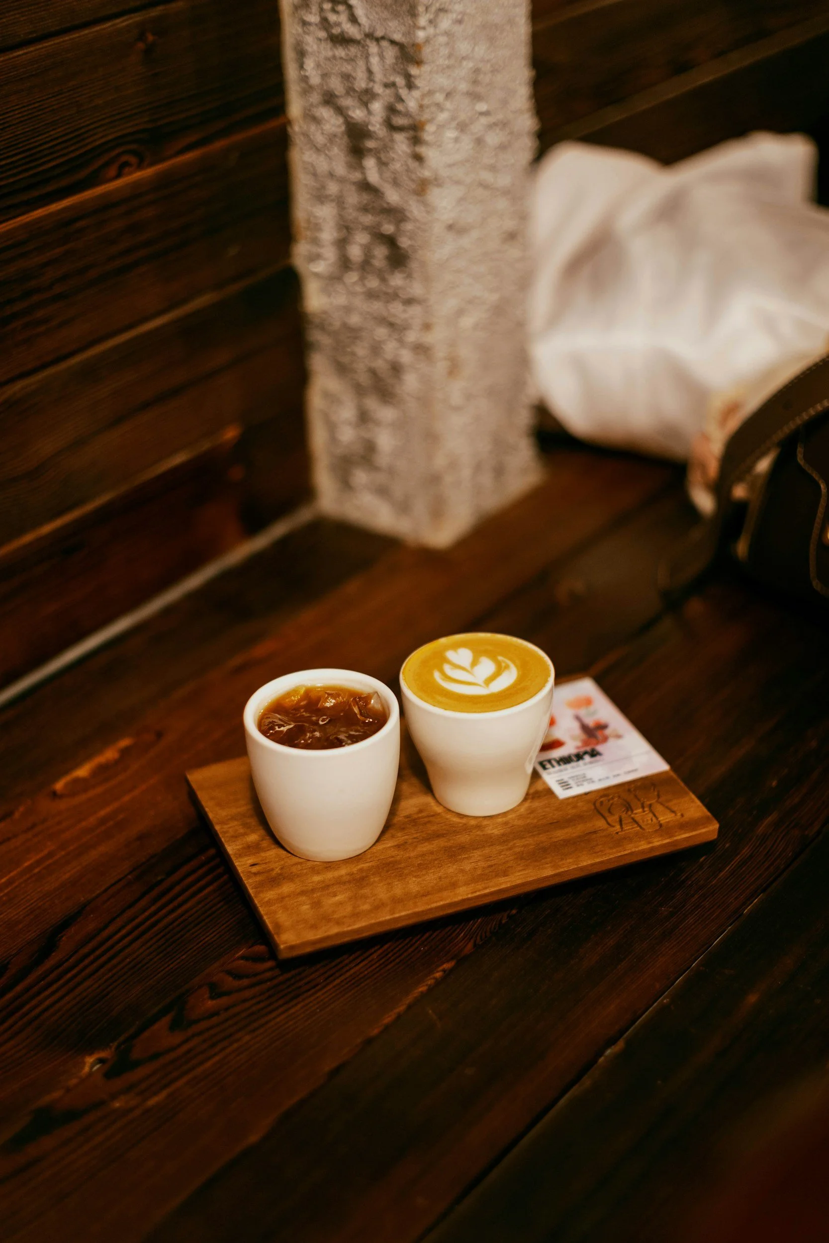 Two white cups of coffee on a wooden serving board on a dark wooden table, with a latte art and iced coffee, in a cozy cafe setting.