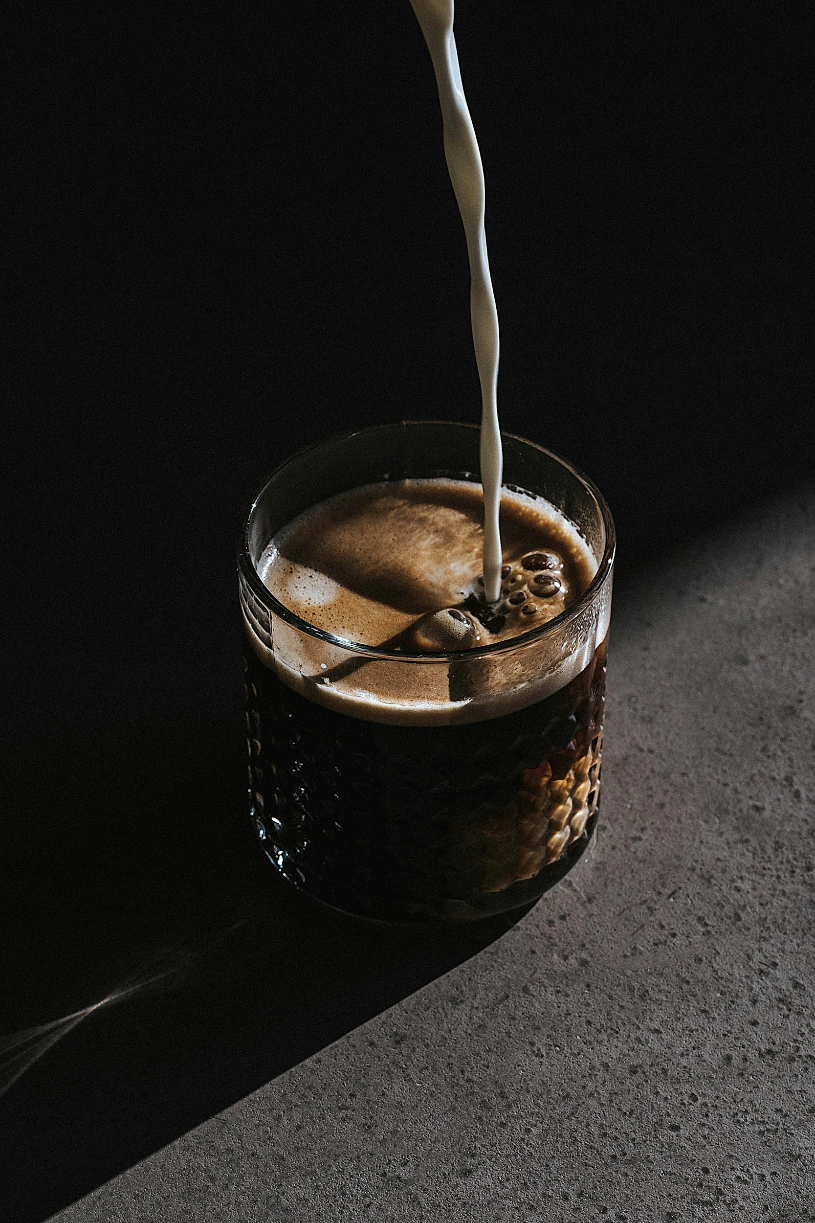 A glass of dark cola with ice cubes, with milk being poured into it in a dimly lit setting.
