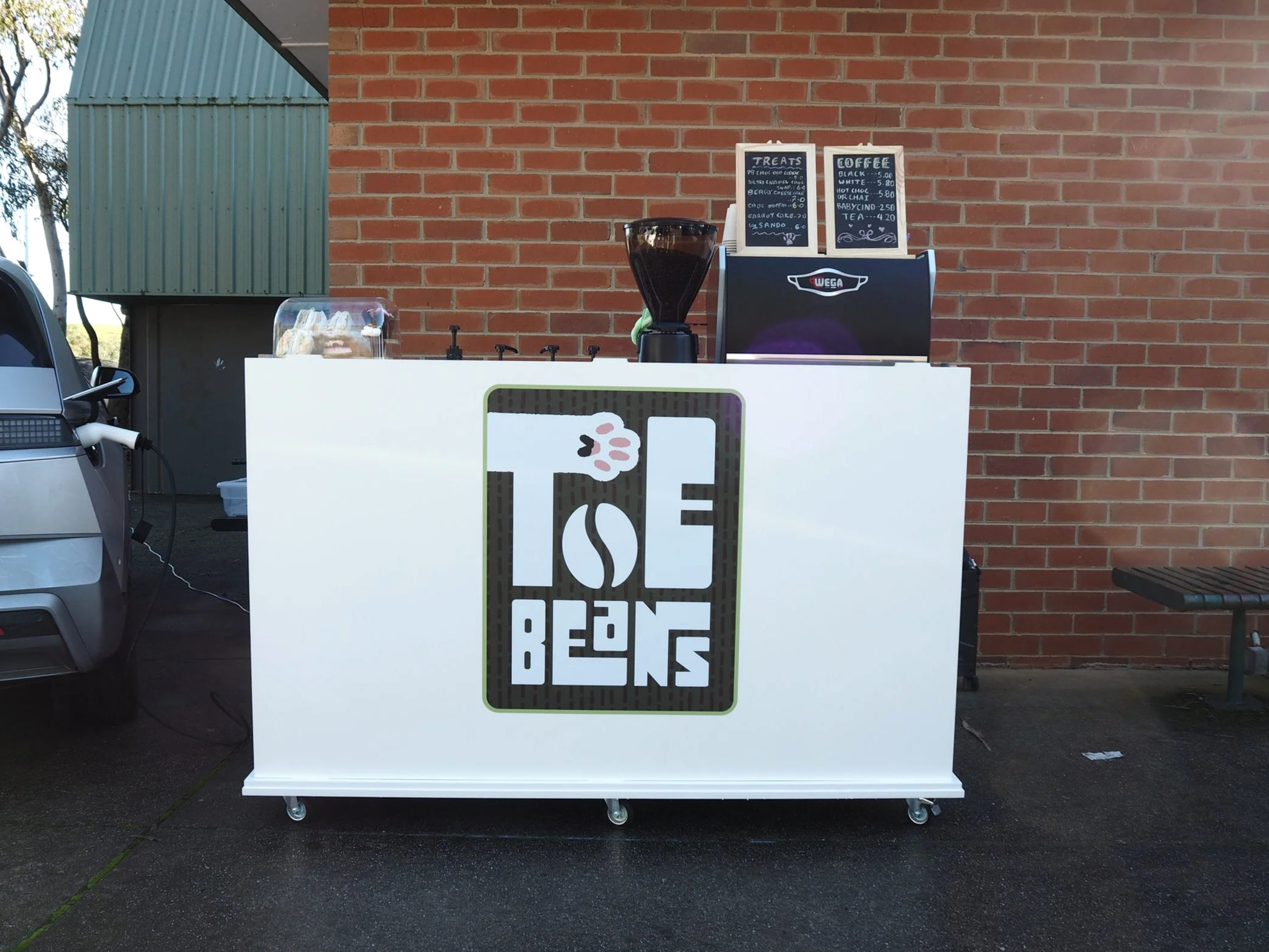 White event cart serving coffee with a coffee machine, grinder and food display. A logo sticker with Toe Beans is displayed on the front of the cart.