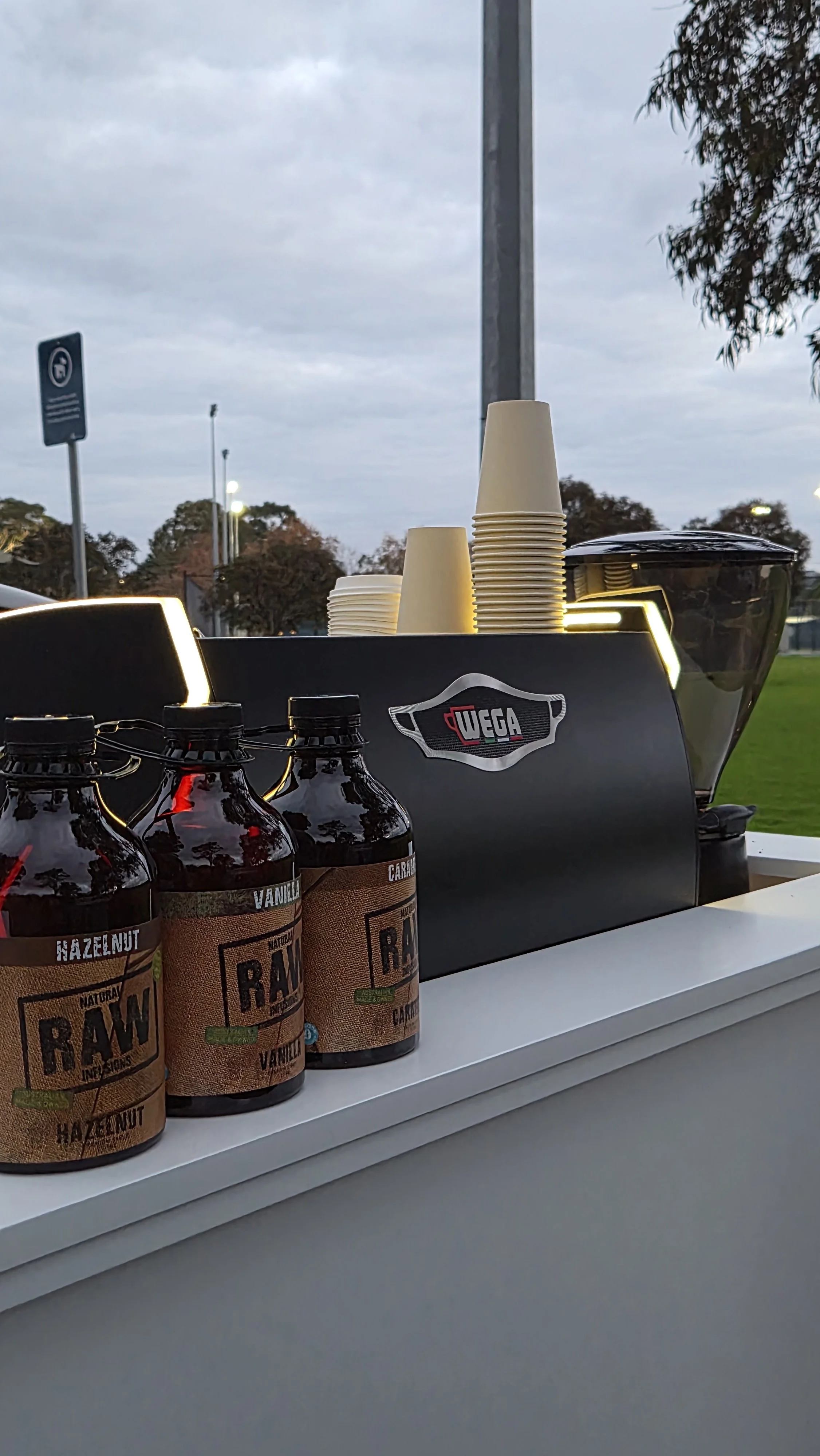 Coffee Syrup bottles labeled 'RAW'. Cups and a coffee machine on a cart outdoors in cloudy weather.