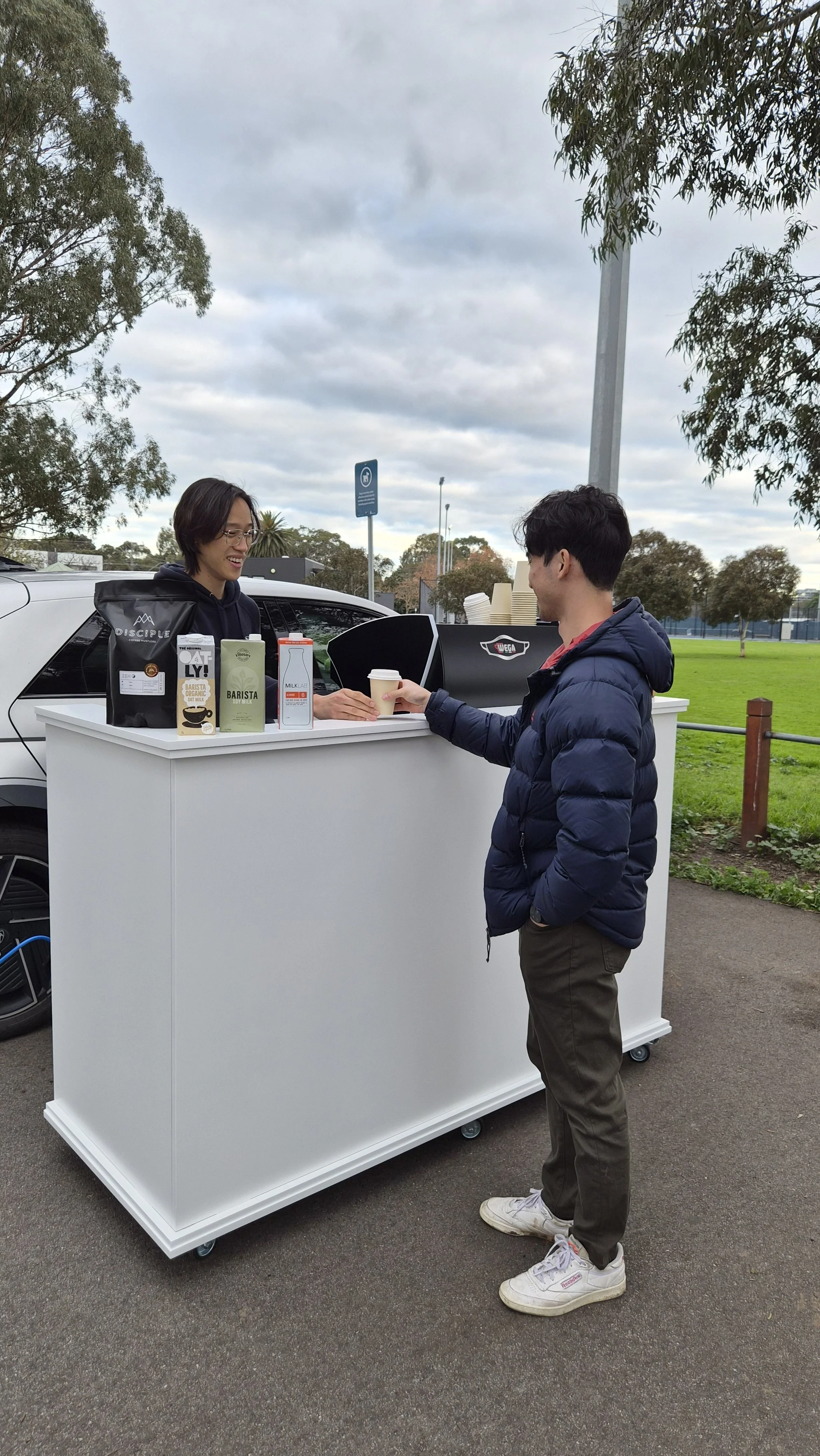 A person in a puffy jacket receives a coffee from a barista at a white mobile coffee stand outdoors during the day. The barista is smiling and wearing glasses, and there are various coffee cartons and a coffee machine on the stand. The background sho