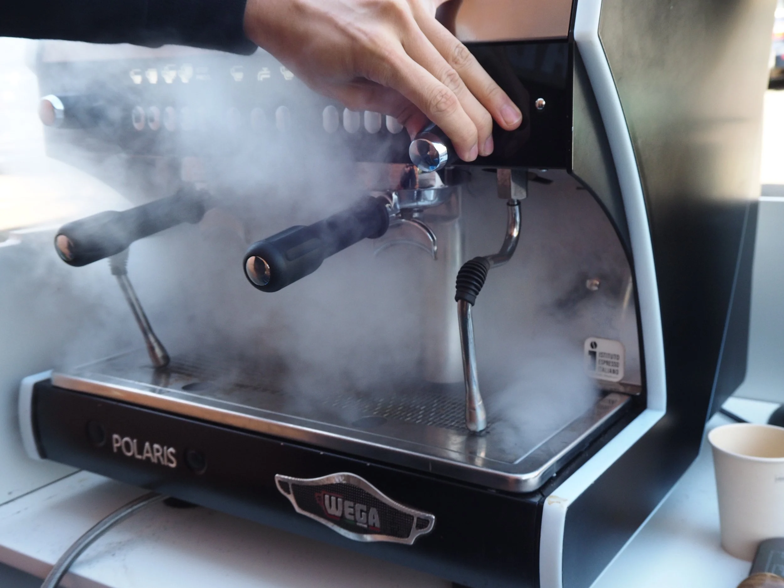 Close-up of a hand adjusting a knob on a Polaris Wega coffee machine with steam rising from it.
