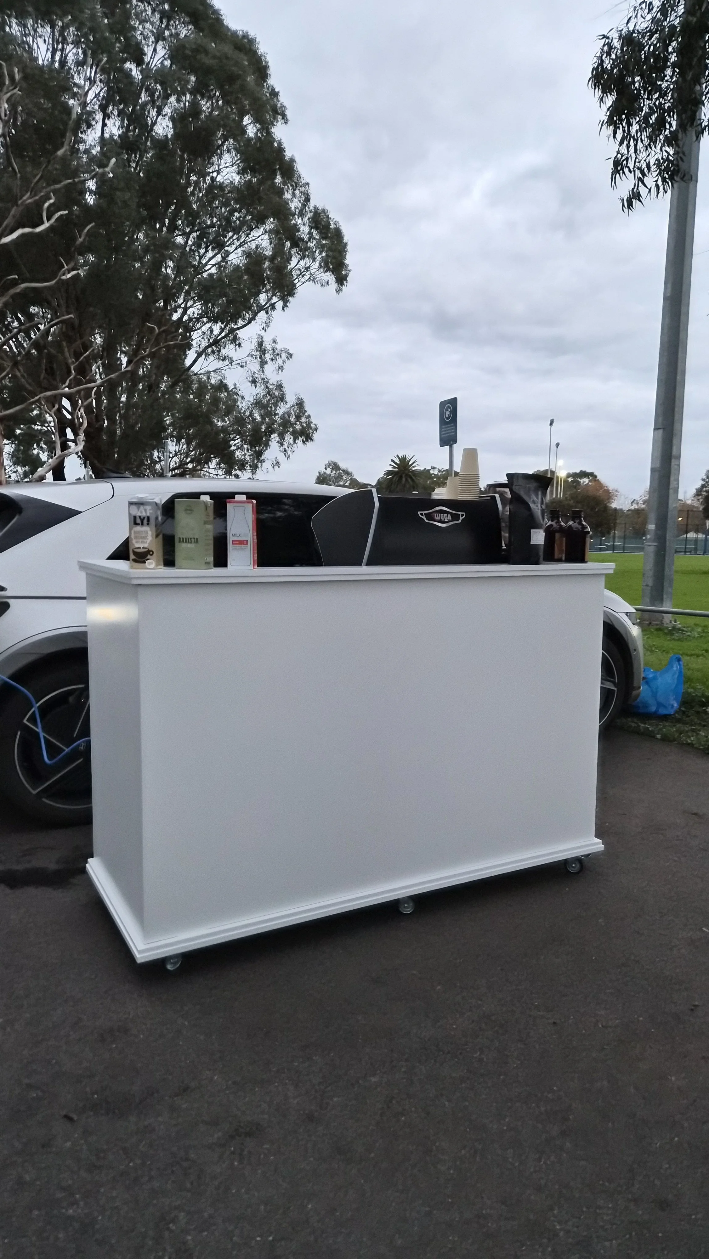 A white mobile coffee stand on wheels with various coffee cartons and equipment on top, parked outdoors with trees and a cloudy sky in the background.