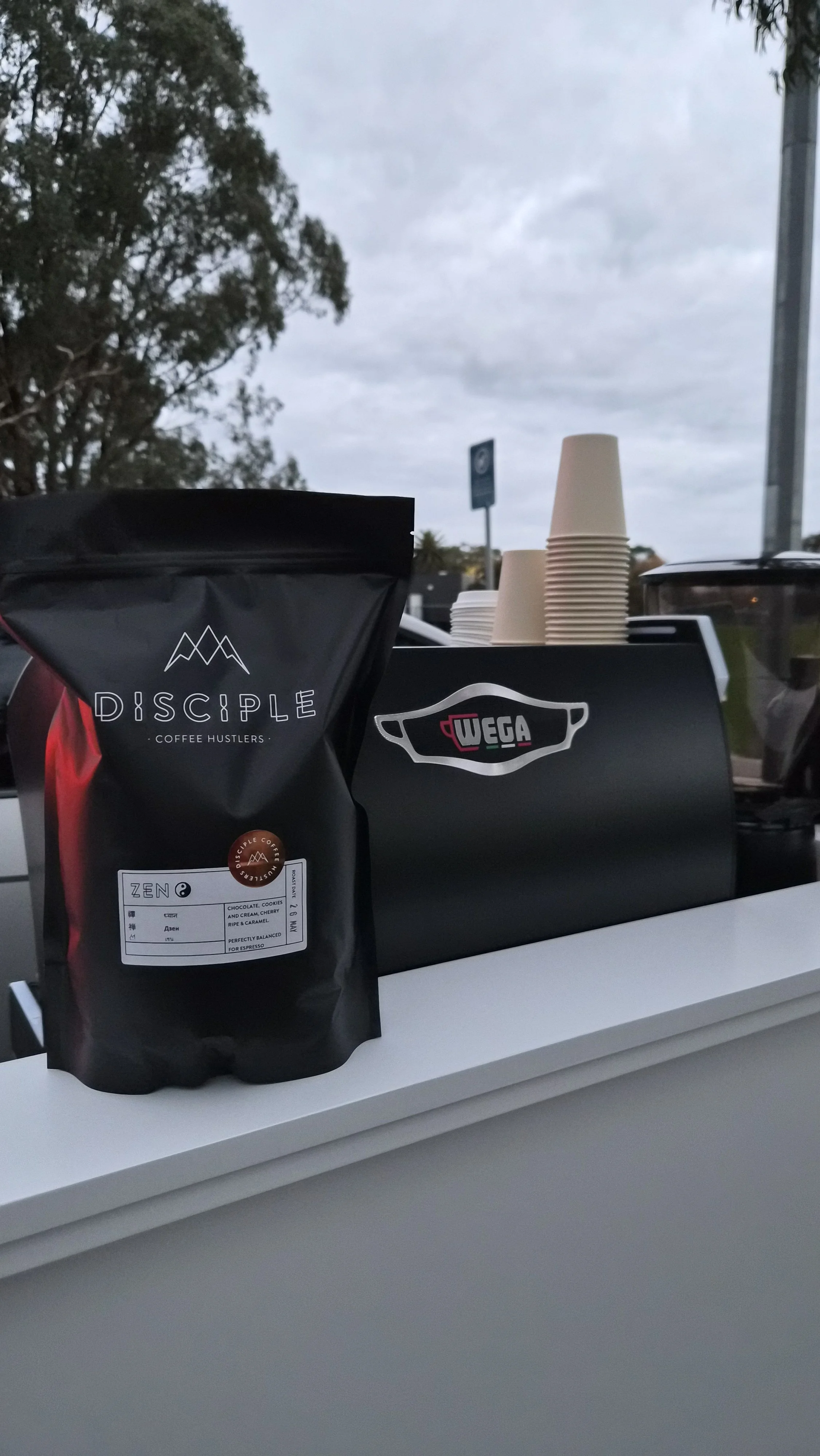 A black bag of coffee labeled 'Disciple Coffee Roasters' on a white counter with disposable coffee cups and a black box in the background, outdoor setting with trees and cloudy sky.