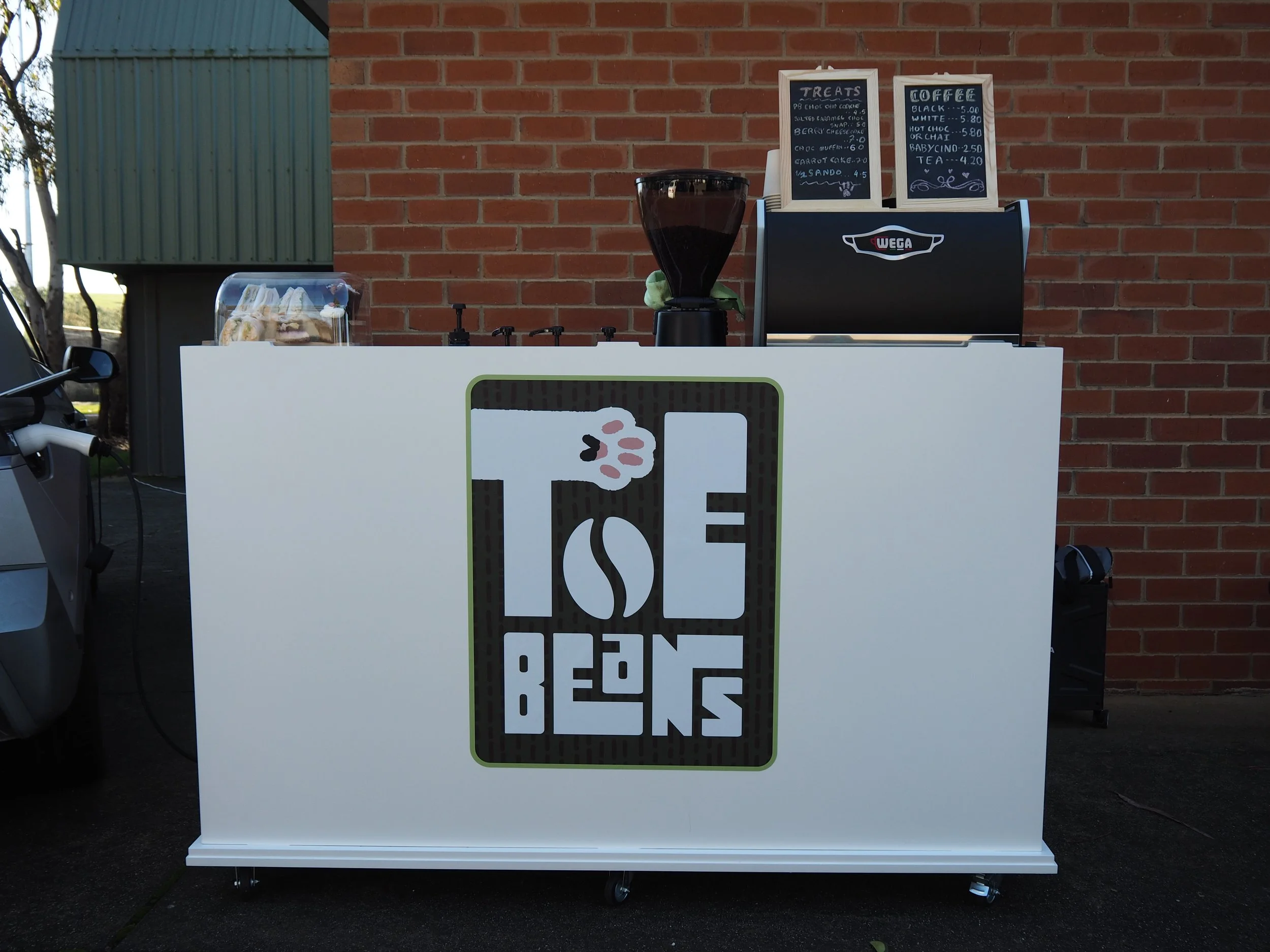 Outdoor ice cream stand with a white front panel that has a logo and the words 'Ice Creams' on it; behind the stand, against a red brick wall, are two chalkboards listing treats and drinks, a large black grinder or blender, and a transparent containe