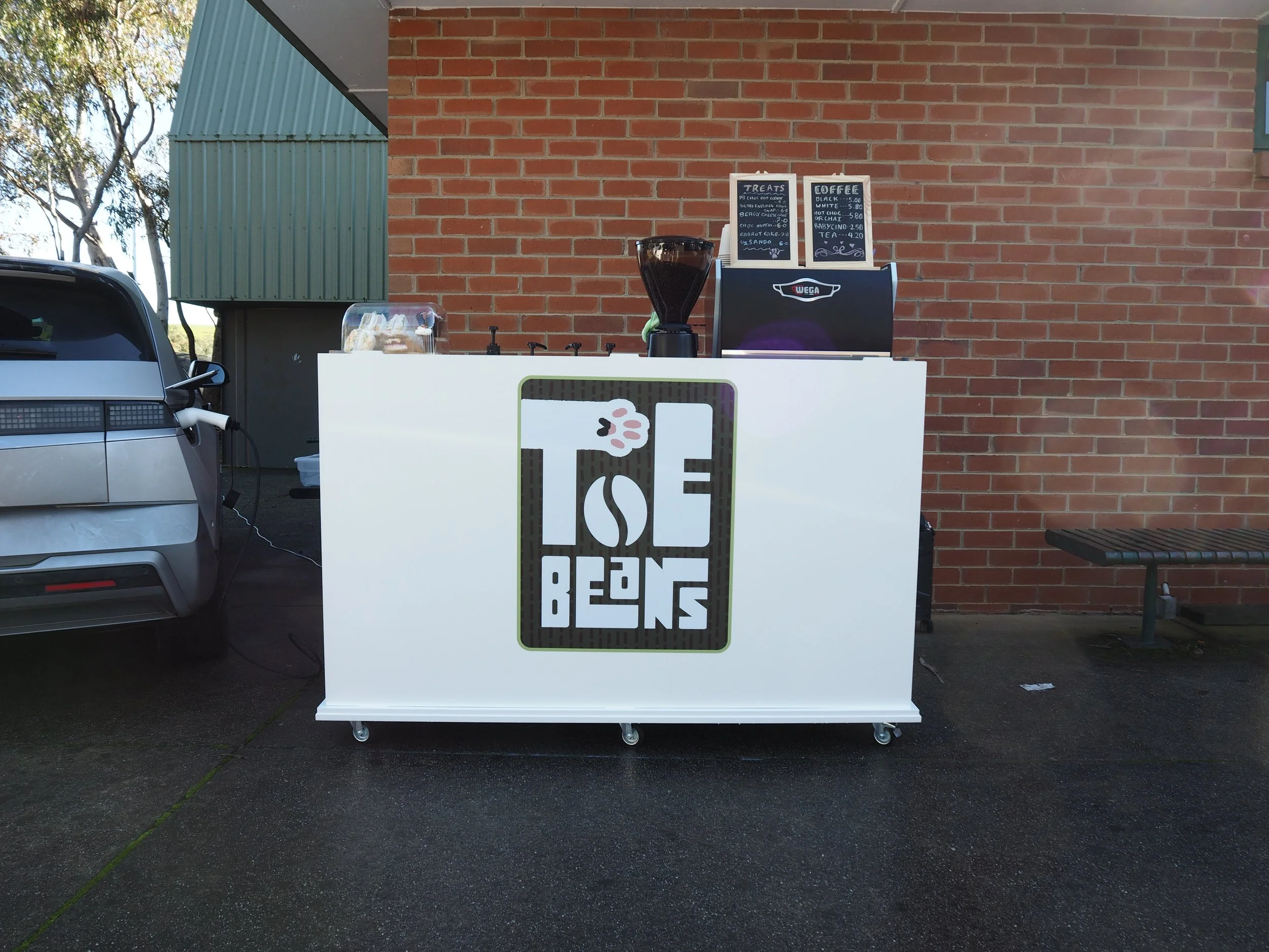 Food truck with a sign reading 'Lost Beans' parked outside a building with a brick wall. The truck offers treats like cookies and coffee, with a menu displayed on a chalkboard. Part of a silver car is visible in the foreground.