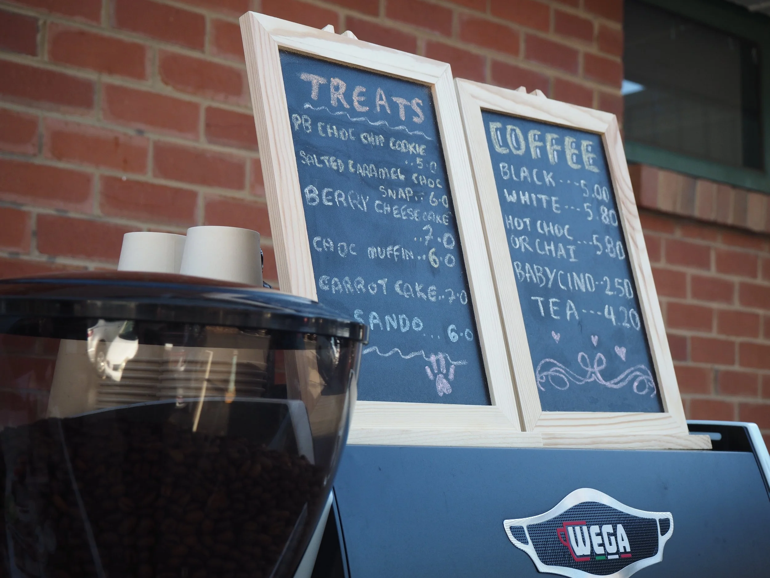 A chalkboard menu displaying items such as treats and coffee, with a brick wall in the background and a storage container with coffee beans in the foreground.