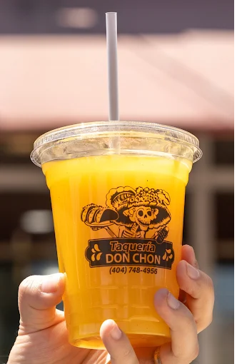 A hand holding a plastic cup filled with yellow mango smoothie with a straw, featuring a taqueria logo.