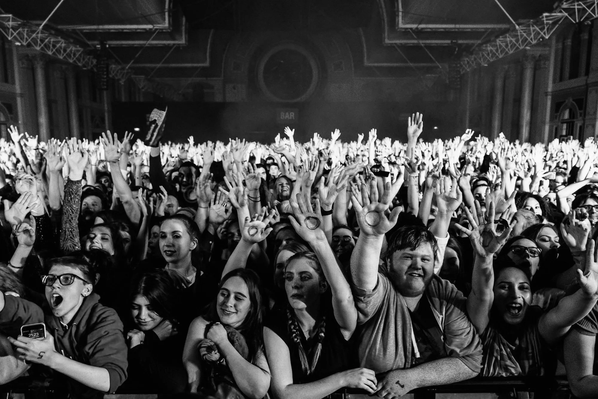 A large crowd of people at a concert or event, raising their hands and enjoying the moment inside a spacious indoor venue with high ceilings and scaffolding.