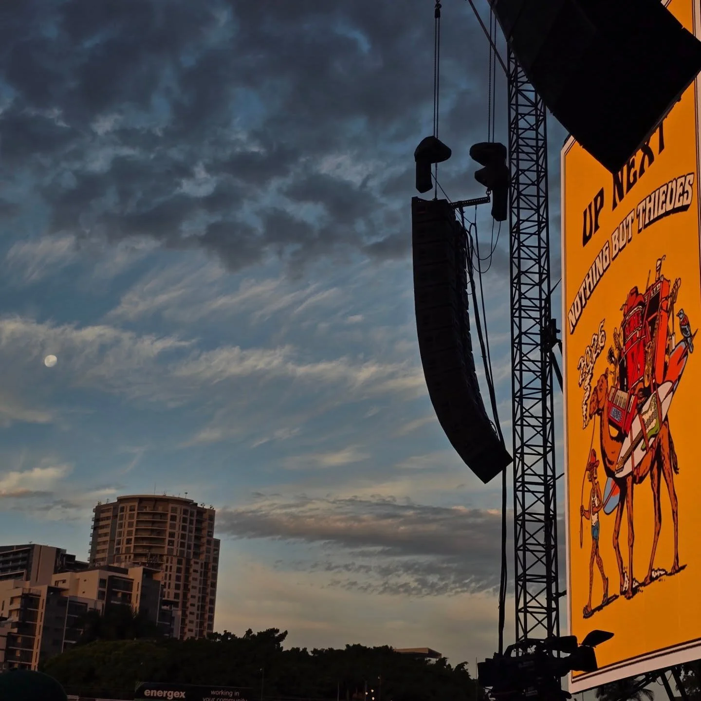 An outdoor stage setup with a large yellow banner featuring a cartoon illustration of a person riding a camel and playing a drum, with the text "UP NEXT" written above. The sky is partly cloudy with a visible moon and some high-rise buildings in the background.