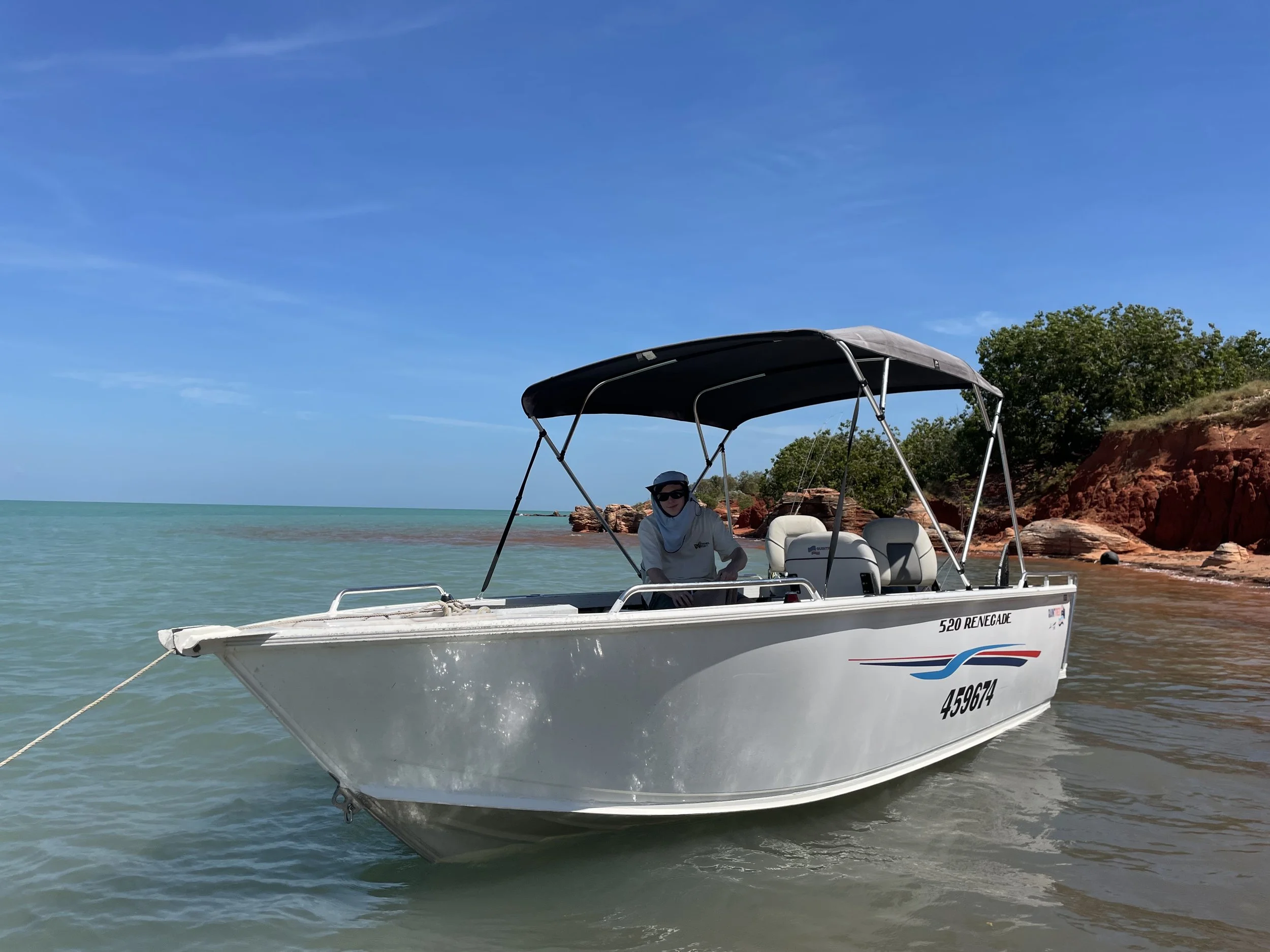 White open fishing boat in the foreground, equipped with 4 seats and shaded canopy. Man sitting under the shade with fishing gear on. Boat sits on clear blue waters with red cliffs to the right with greenery on top.