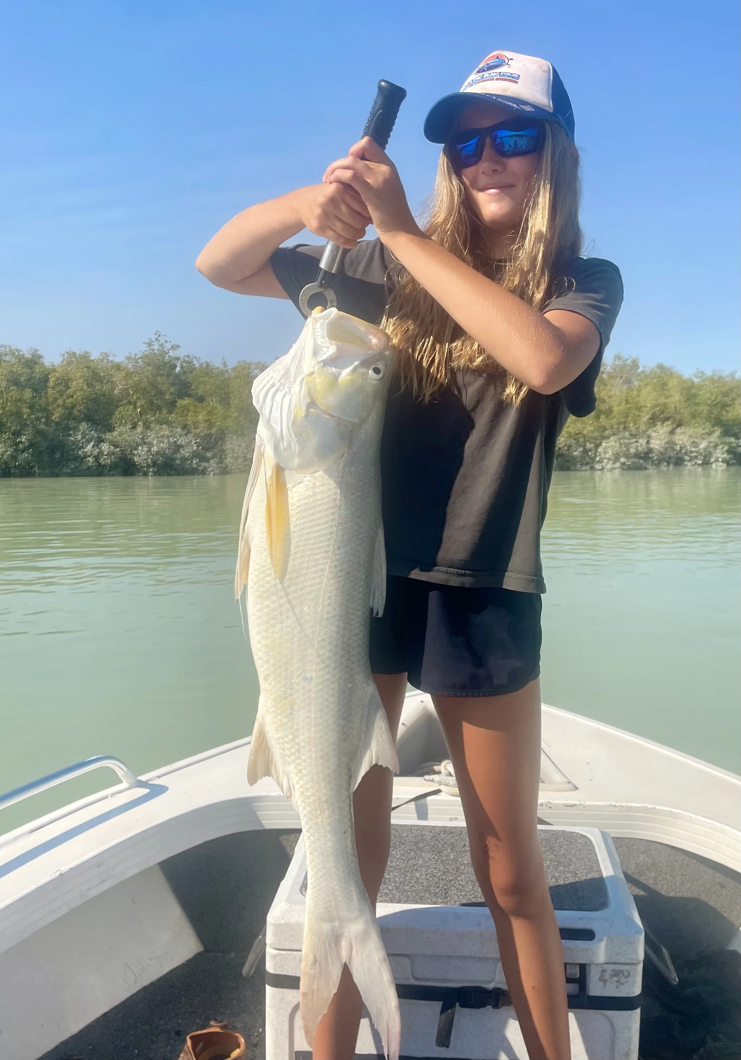 Girl standing in fishing boat holding up large threadfin salmon, almost her height. Background displays the creek of the mangroves.