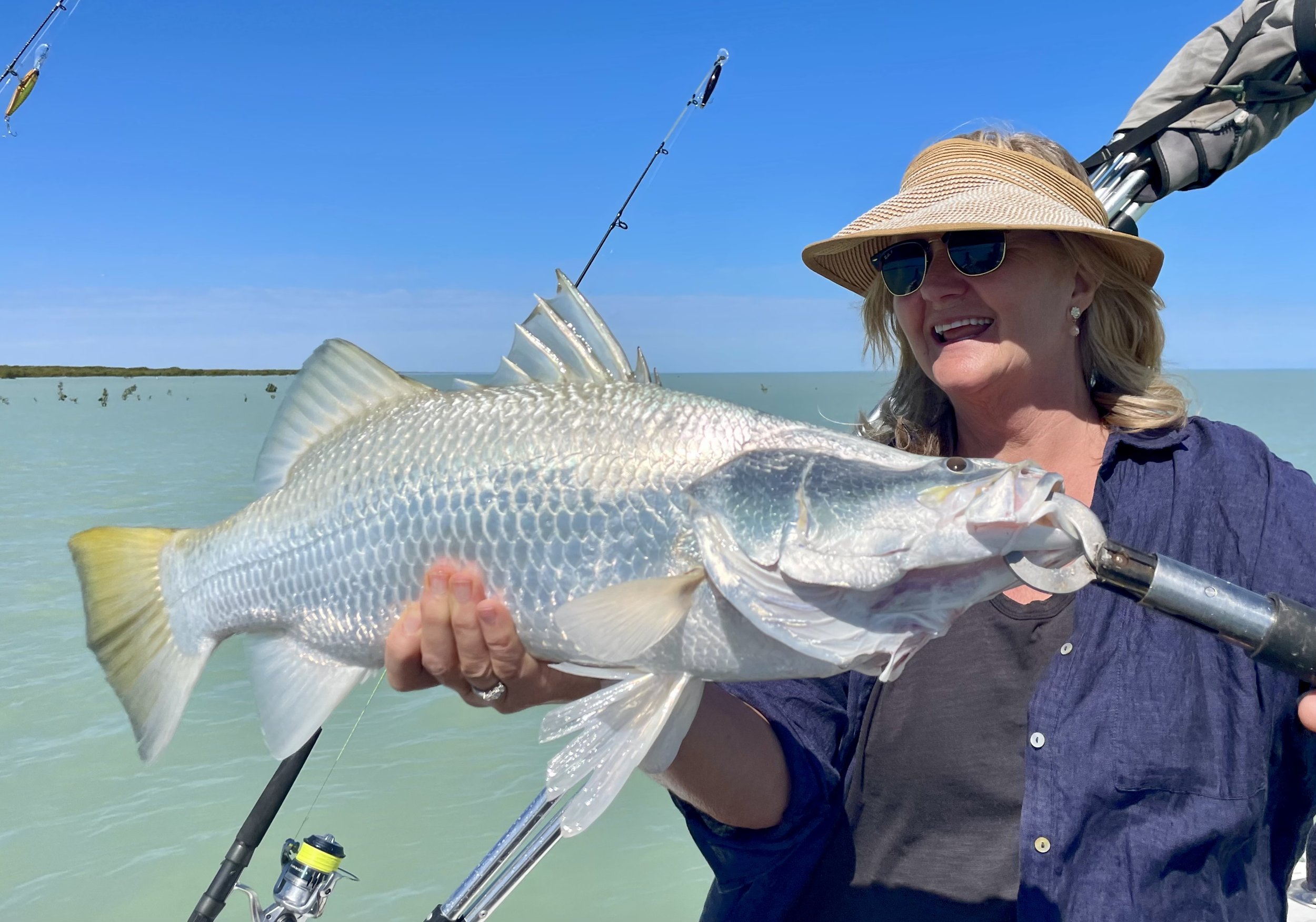 Lady sitting in fishing boat, smiling while holding barramundi. Waters of the creek in the background with clear blue sky.