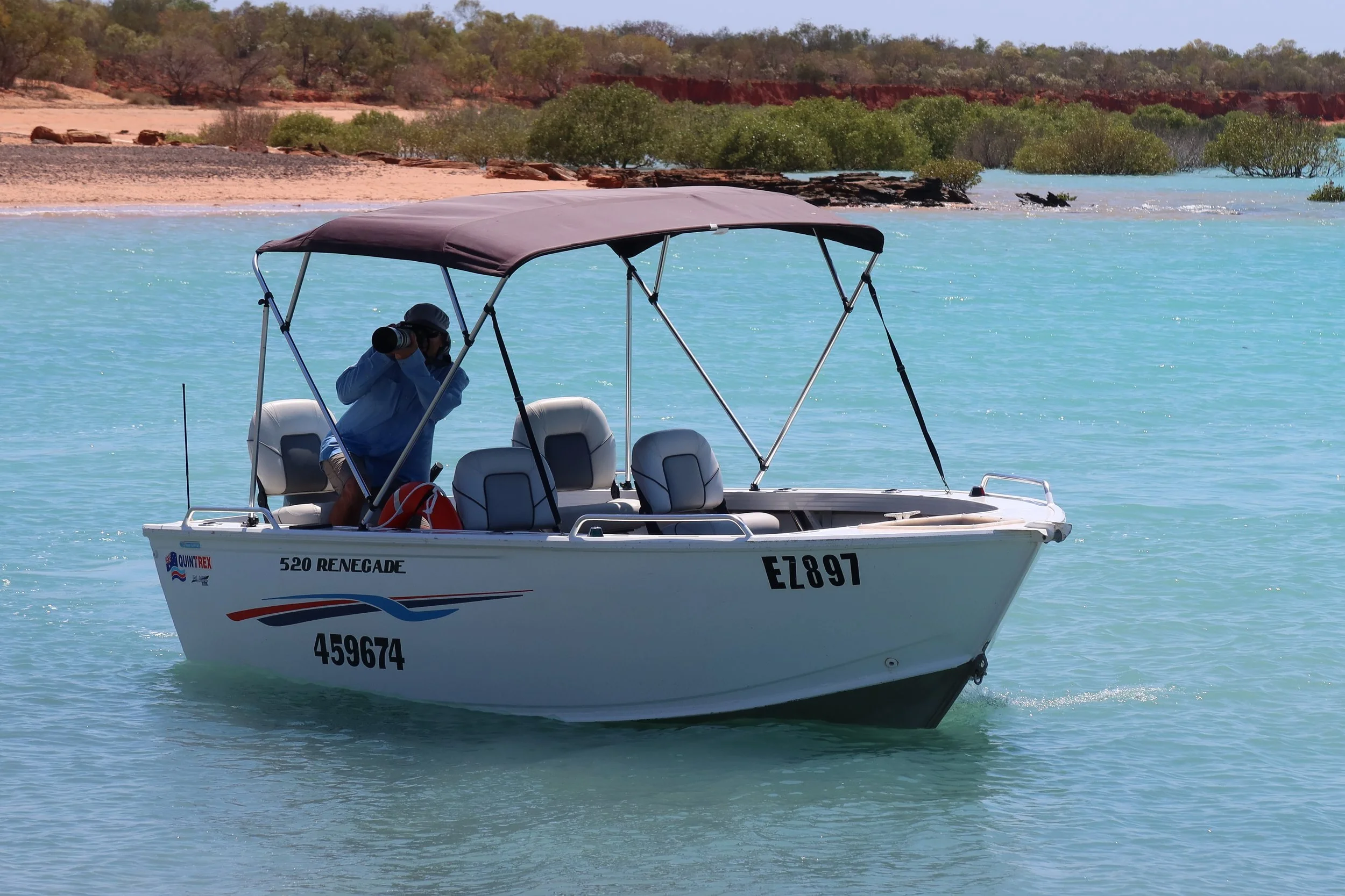 Landscape image of open fishing boat with shaded canopy. Boat sits on blue ocean waters with beach and mangroves in the background. 4 Chairs visible in the boat with fisherman standing up to take photos of wildlife.