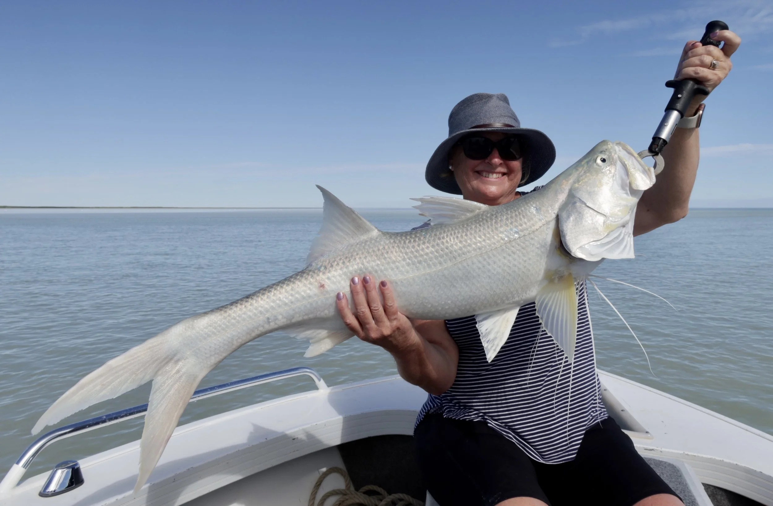 Lady sitting in a white boat holding up huge threadfin salmon. View of the ocean and clear blue skies in the background.