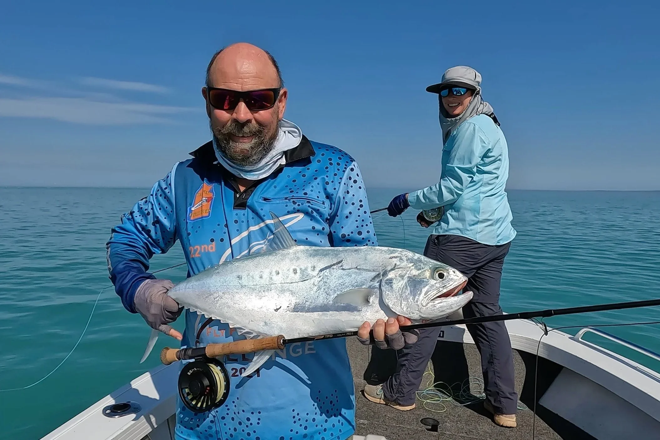 A couple standing in a boat on the water holding Tuna and fly fishing rod.