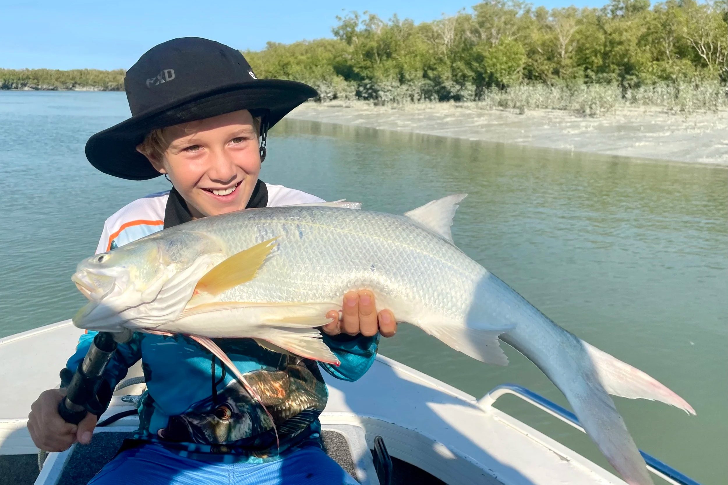 Boy in fishing attire on a boat holding threadfin salmon. Broome bay in the background.