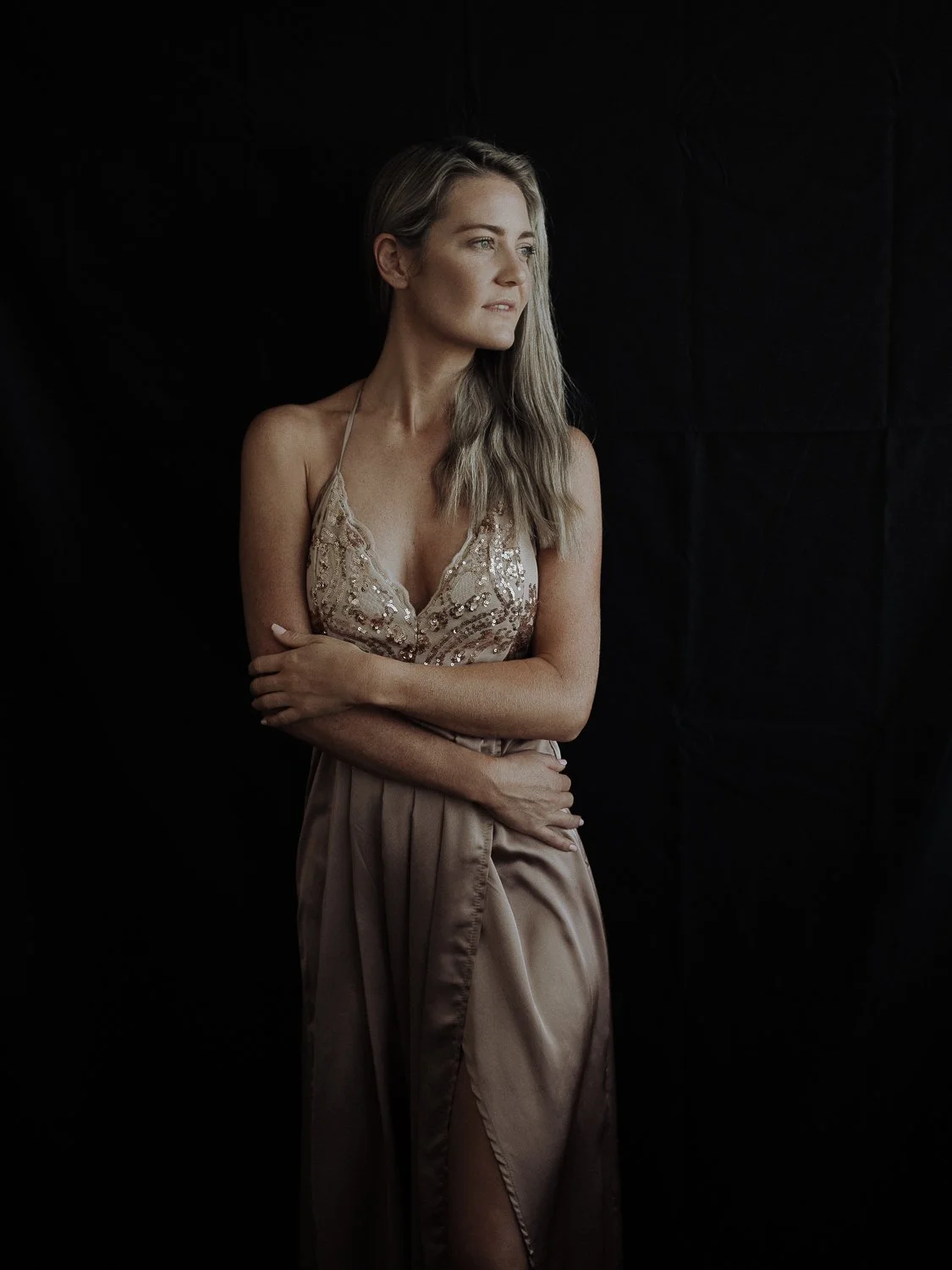 A woman with long blonde hair wearing a beige, sequined dress stands against a dark background, looking to her right with arms crossed.