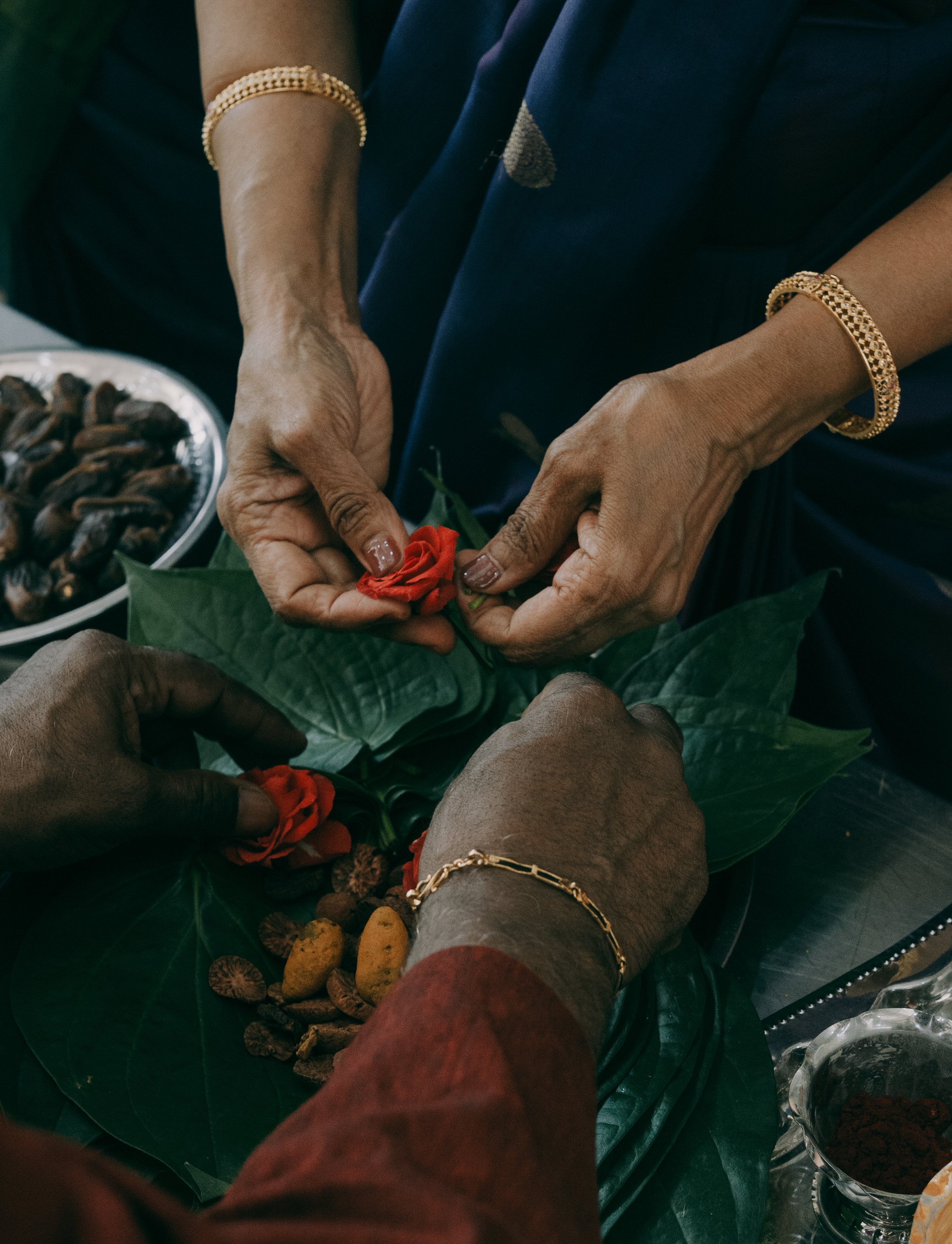 Multiple people are gathering around, exchanging a red flower, possibly a rose, in a setting with food and snacks visible in the background, including a plate of dried snacks and bowls of what appear to be dried fruits or nuts.