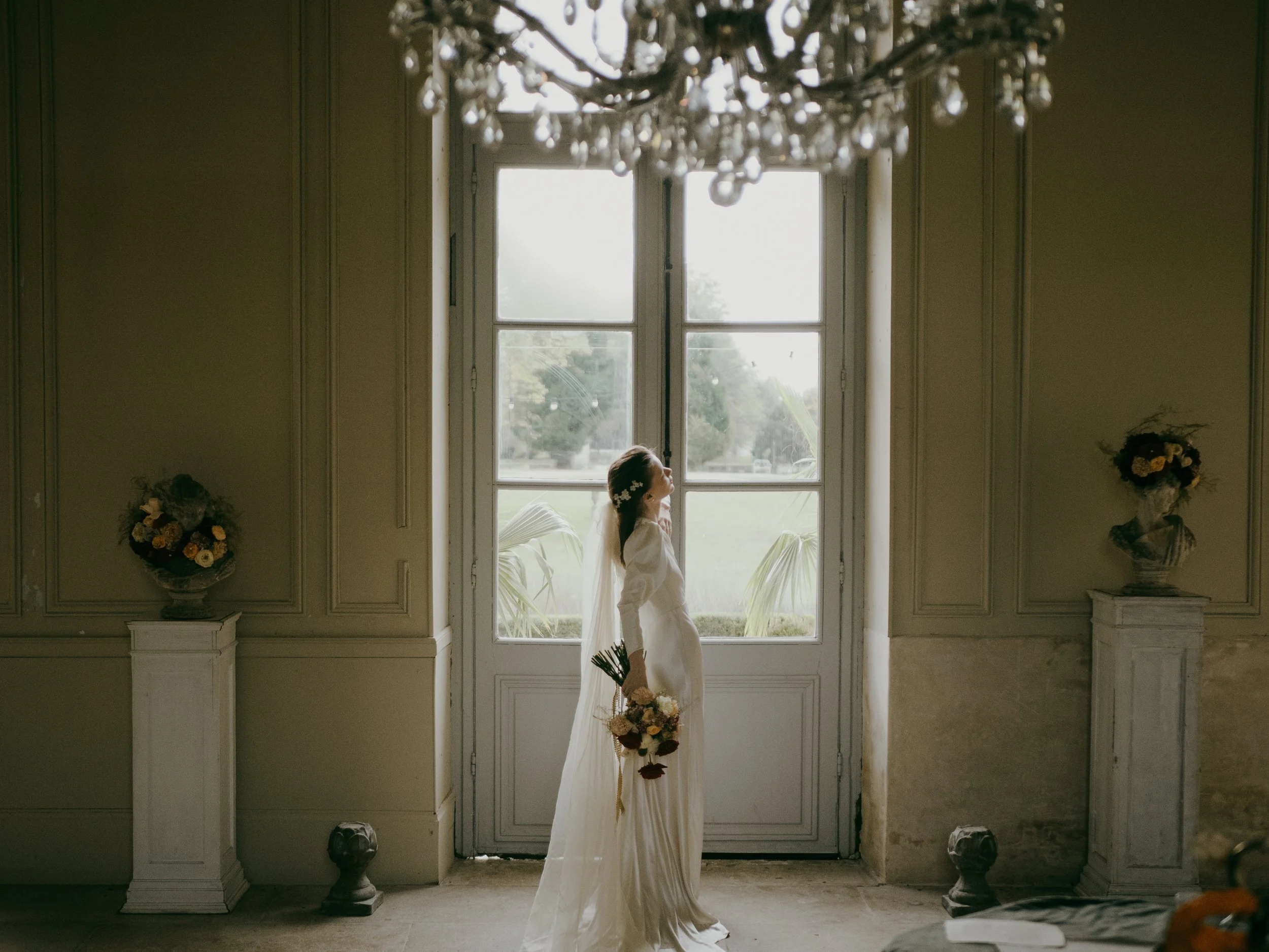 A bride in a white dress holding a bouquet of flowers, standing in front of large windows with curtain panels, with a chandelier overhead and floral arrangements on either side of the window.
